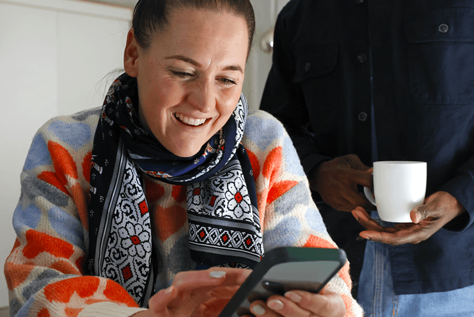 A person seated indoors looks at a smartphone and interacts with the screen. Another person stands nearby holding a white mug, creating a relaxed home environment.